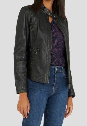Femme portant une veste en cuir noire sur un haut sombre à motifs et un jean bleu sur fond uni.
