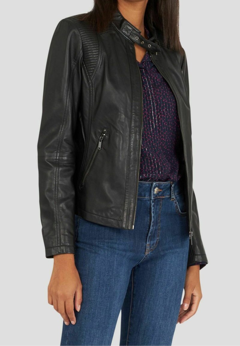 Femme portant une veste en cuir noire sur un haut sombre à motifs et un jean bleu sur fond uni.