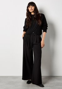 Czarny sweter w splot kablowy z cropped designem, w połączeniu z czarnymi spodniami o wysokim stanem i szerokich nogawkach. Spodnie mają wiązanie w pasie oraz kieszenie boczne.