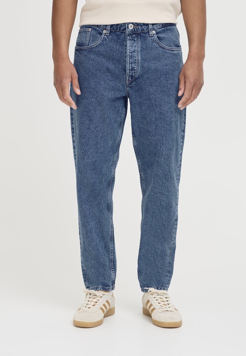Jean en denim bleu avec une coupe droite, cinq poches, fermeture par bouton et zip, et une texture délavée. Porté avec des baskets de couleur claire.