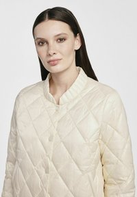 Kremowa pikowana kurtka z wysokim kołnierzem i zapięciem na guziki, z diamentowym wzorem i gładką, błyszczącą teksturą.