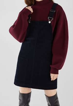 Kobieta w czarnej sukience na szelkach z sztruksu z przednimi kieszeniami, bordowym golfem z długim rękawem i czarnych kozakach za kolano.