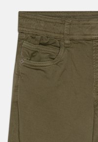 Olivgrön bomullshakama med femfickorsdesign, texturerat tyg och sömnadsdetaljer. Inkluderar en liten dekorativ ficka.