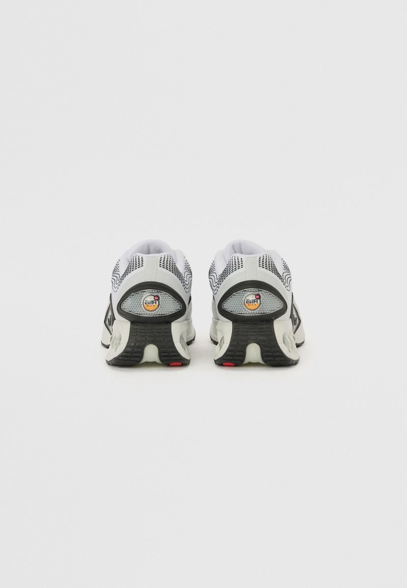 Des chaussures de course blanches et noires avec une tige en maille, un col rembourré, un coussin d'air visible au talon et une semelle en caoutchouc texturée avec des accents rouges.