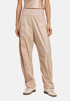 Losse beige trainingsbroek met elastische tailleband en zijstrepen, gedragen met beige en witte sneakers op een staand model.