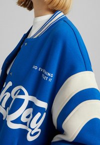 Modrá varsity bunda s bílými pruhy, se zapínáním na přední tlakové knoflíky a nápisem "DĚLEJ COKOLI." Měkká textura, sportovní design.