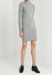 Abito grigio a maniche lunghe con silhouette aderente e scollatura a barchetta, decorato con dettagli al colletto, abbinato a scarpe nere lucide.