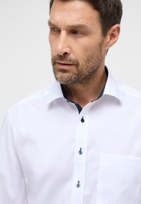 Biała koszula na guziki o dopasowanym fasonie, z granatowym wzorem w kropki pod kołnierzykiem i niebieskimi guzikami. Z przodu znajduje się jedna kieszonka na piersi.