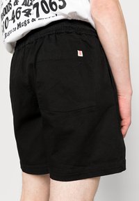 Black cotton shorts with an elastic waistband, featuring a back pocket and a small red label on the left side. Textured fabric.