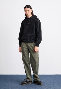 Crna pamučna hoodie sa vezicama, s kenguruvim džepom. Uparen s svijetlozelenim širokim hlačama i crnim platformama.