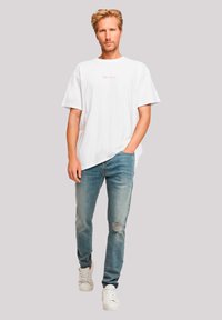 Witte katoenen t-shirt met een kleine rode print op de voorkant, gecombineerd met lichtblauwe versleten jeans en witte sneakers, staande voor een neutrale achtergrond.