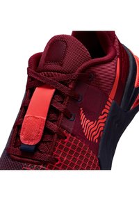 Gros plan d'une chaussure de sport rouge avec un mesh texturé, des lacets rouges, une bride rouge et un motif rouge ondulé sur la partie latérale.