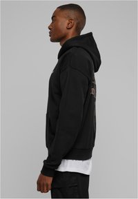 Sudadera con capucha negra con un bolsillo, que presenta una textura suave, puños acanalados y una gran impresión gráfica en la parte trasera. Se lleva sobre una camisa blanca.