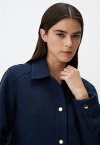 Marineblauer Wollmischjacke mit kragenförmigem Ausschnitt, goldenen Druckknöpfen und seitlichen Taschen. Glatte Textur mit strukturierten Nähten und Bündchen.