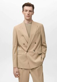 Blazer beige doppiopetto con rever a punta, due tasche frontali e bottoni marroni, abbinato a un top in maglia a coste beige e pantaloni coordinati.
