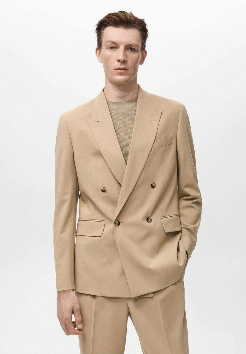 Blazer beige doppiopetto con rever a punta, due tasche frontali e bottoni marroni, abbinato a un top in maglia a coste beige e pantaloni coordinati.