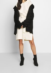 Cárdigan negro de textura grande sobre un vestido beige con cinturilla ajustada. Botas de tacón hasta el tobillo negras completan el conjunto.