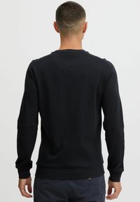 Sweatshirt noir à manches longues en tissu texturé, avec des poignets et un ourlet côtelés, présentant un col rond et un détail de couture au dos.