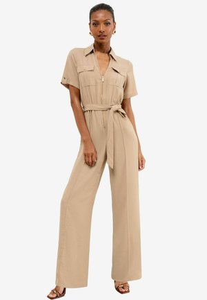 Vrouw poseert in een beige korte mouw jumpsuit met rits aan de voorkant, borstzakken, strikceintuur, brede benen en open teen hakken.
