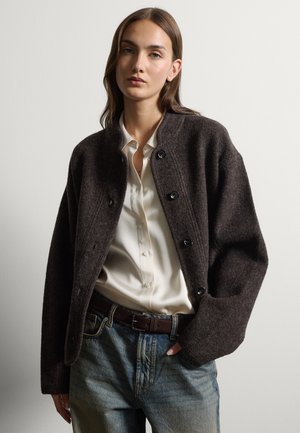 Frau mit langen braunen Haaren, die eine dunkle, geknöpfte Wolljacke über einem weißen Hemd und blauen Jeans trägt, steht vor einem einfarbigen hellen Hintergrund.