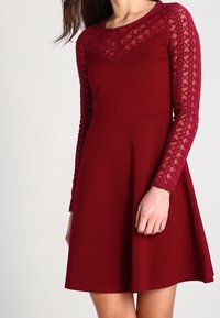 Robe bordeaux avec manches longues en dentelle, corsage ajusté et jupe évasée. La dentelle présente un motif géométrique le long des bras.