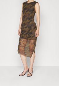 Vestido sin mangas color verde oliva con un sobrecuerpo transparente, que presenta un patrón geométrico y floral, y una abertura lateral. Complementado con sandalias negras con tiras.