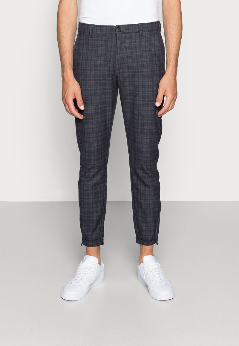 Chinos van donkere geruite stof, met een aansluitend ontwerp, enkellang, minimale ritsaccenten bij de manchetten, gecombineerd met witte sneakers.