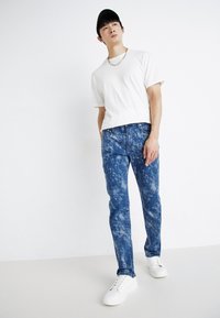 Lett og lett hvit t-skjorte, blå tie-dye jeans i forskjellige nyanser, og hvite joggesko; person med svart caps og sølv-halskjede.