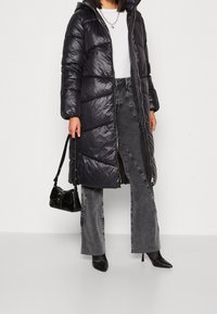 Long manteau gonflé en matériau noir brillant avec capuche, doté d'une fermeture éclair à l'avant, avec un matelassage à motifs, porté sur un jean gris à jambes larges et des talons noirs.