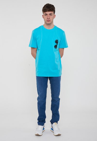 Maglietta turchese con grafica di occhiali da sole neri sulla tasca, abbinata a jeans blu e scarpe bianche. Design semplice, a maniche corte.