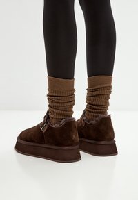 Bruine suède enkelboots met een dikke plateauzool, voorzien van een gespdetail en een zachte voering, gecombineerd met geribde bruine beenwarmers.