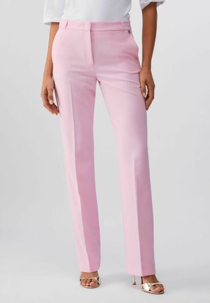 Kobieta ubrana w proste, jasnoróżowe spodnie i biały top oraz złote sandały na wysokim obcasie z odkrytymi palcami, stojąca na jednolitym tle.