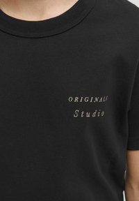 Černé bavlněné tričko s krátkým rukávem, se svělým béžovým textem, který zní "ORIGINALS" nad "Studio" na levé části hrudníku.