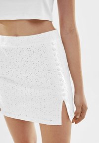 Vit minic kjol med blommiga utskärningar, en sidoklyvning och dekorativa knappar längs den vänstra sömmen. Tillverkad av lätt tyg.