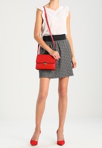 Rote Handtasche mit Klappe und goldener Schnalle, kombiniert mit einem weißen Oberteil mit Spitze und einem schwarz gemusterten Rock. Rote High Heels runden das Outfit ab.