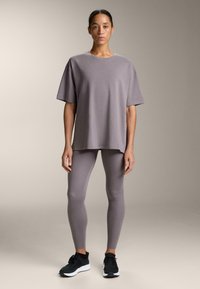 Svetlosivé oversize tričko s okrúhlym výstrihom, kombinované s priliehavými svetlosivými legínami a čiernymi športovými topánkami, na tlmenom pozadí.