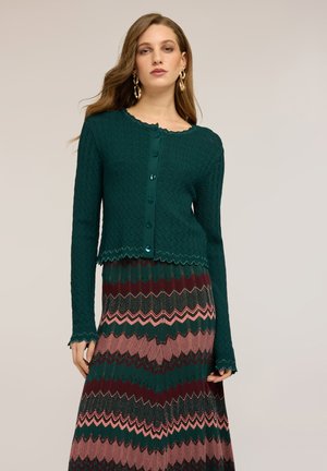 Kobieta z długimi włosami ubrana w teksturowany miętowy kardigan na guziki oraz wielokolorową spódnicę w zygzakowaty wzór na jednolitym jasnym tle.