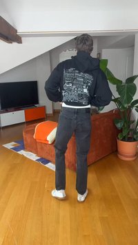 Άτομο που στέκεται με την πλάτη στην κάμερα, φορώντας μαύρο φούτερ με λευκό σχέδιο, σκούρο τζιν και λευκά αθλητικά παπούτσια σε σαλόνι με πορτοκαλί καναπέ.