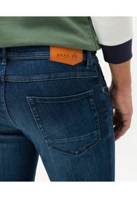Dark blue denim jeans with a smooth texture, featuring a brown leather waistband label with "BRAX" and a decorative stitching detail on the pocket.
