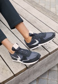 Mörkgrå sneakers med mesh- och mocka-material, med en vit Nike-logga, svarta snören och en vit hälflik. Platt gummisula.