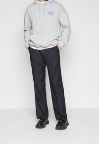 Grå huvtröja med grafik på bröstet och ärmarna, i kombination med svarta kostymbyxor och svarta skor, mot en vit bakgrund.