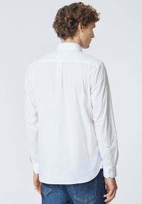 Camicia bianca a maniche lunghe con colletto standard, dettaglio plissé sul retro e polsini con bottoni. Realizzata in tessuto liscio, abbinata a jeans blu.