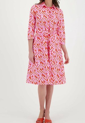 Mujer con vestido rosa hasta la rodilla con patrón geométrico rojo y naranja, mangas tres cuartos y lazo en la cintura, combinado con tacones rojos de punta abierta.