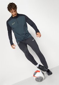 Mannen sportoutfit met een donkergroen sweater met ritssluiting en lange zwarte mouwen, gecombineerd met strakzittende zwarte broek en zwarte voetbalschoenen.