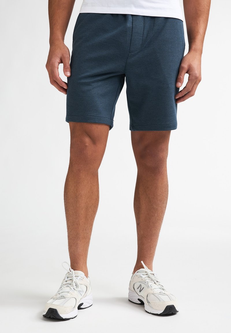 Pantalones cortos deportivos de color azul oscuro hechos de tela texturizada, con una cinturilla elástica y un diseño hasta la rodilla, combinados con zapatillas blancas para correr.