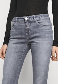 Femme portant un jean slim gris clair et un haut à manches longues noir, les mains détendues le long du corps, la taille et le haut des cuisses visibles.