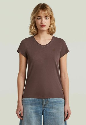 Femme aux cheveux blonds mi-longs, portant un t-shirt marron uni à manches courtes et col en V ainsi qu'un jean bleu sur un fond clair.