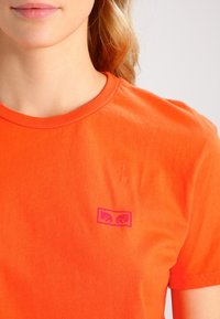 Camiseta de algodón naranja con cuello redondo, que presenta un pequeño logo rosa impreso en el área izquierda del pecho. Textura suave, diseño casual.