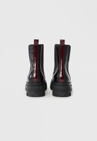 Bottes de cheville en cuir noir texturé crocodile avec une semelle épaisse noire et un panneau arrière laqué bourgogne. Tirants plats pour un enfilage facile.