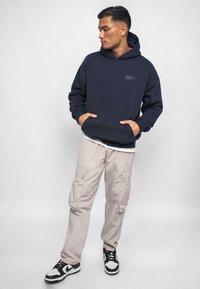 Marineblå hætte sweatshirt med frontlomme, parret med lyse beige cargobukser med lommer og sort-hvide sneakers.
